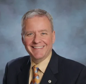 Portrait of a smiling man in a suit and tie against a gray backdrop.
